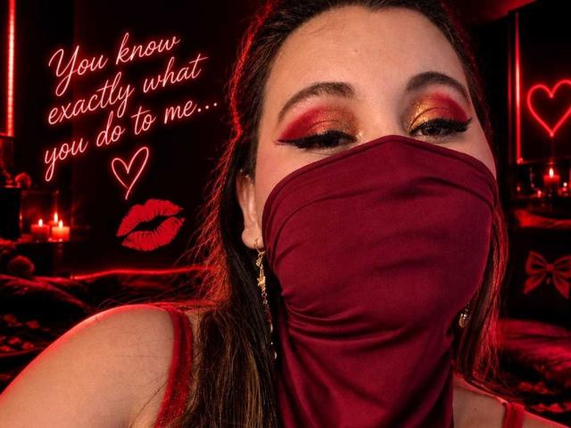 SalmaAzaman with red lipstick, heavy eyeliner, and red eyeshadow, wearing a red mask covering her mouth, and red earrings, looking seductively at the camera. Red background.