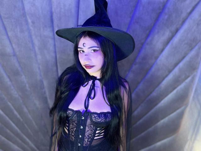 HannahBecker with long black hair, wearing a black lace corset, black witch hat, and dark lipstick, showing her tits, against a fan-like background with blue lighting.