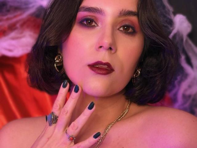 GoddessBar ha i capelli scuri e ondulati, l'eyeliner rosso e il rossetto viola. Indossa orecchini dorati, una collana a catena e smalto per unghie blu scuro. La sua mano tocca il mento..