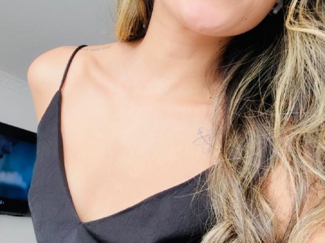 AdharaCooperr's borst met kleine, getatoeëerde tekst aan de rechterkant, draagt een zwarte top met spaghettibandjes, blond haar dat over haar schouder valt, gedeeltelijk gezicht zichtbaar..