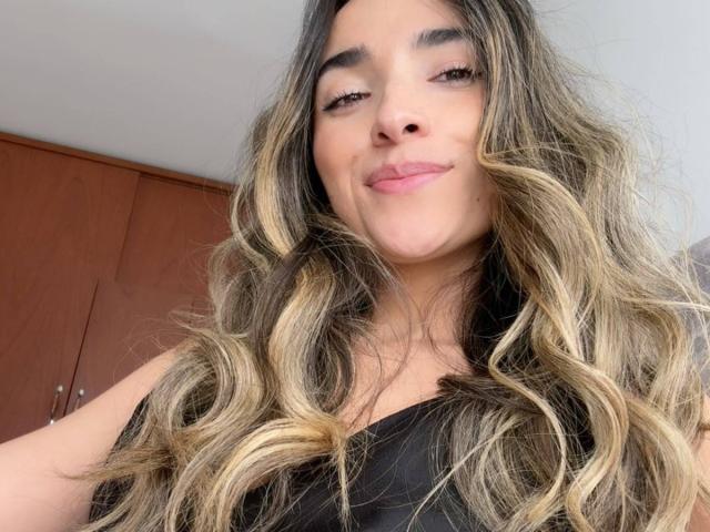AdharaCooperr with wavy, blonde-highlighted brown hair, smiling slightly, pink lips, and wearing a black top, showing her cute face and subtle makeup.