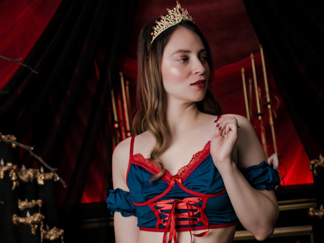 AmberWoody with long brown hair, wearing a gold tiara, blue and red lace-up top, exposing her tits, red lips, and a hand near her chest, in a dark red setting.