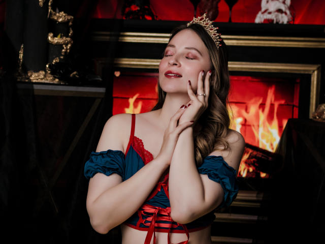 AmberWoody has her eyes closed, smiling with her hands near her face, wearing a red and blue lace-up top with puffed sleeves, and a tiara. Fire is in the background.
