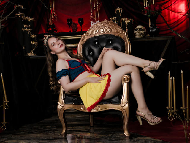 AmberWoody with long brown hair, blue off-shoulder top, yellow skirt with red trim, gold high heels, black gold chair, legs crossed, arms resting, relaxed, red and black background.