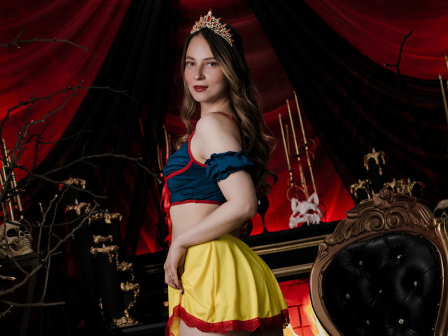 AmberWoody with long brown hair, blue off-shoulder top, yellow skirt, red trim, and gold tiara, showing her ass, standing next to a black chair, red and black backdrop.