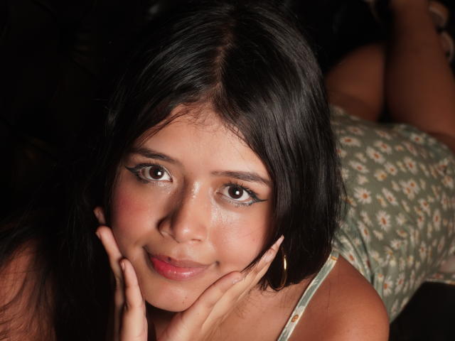 DiamondSweet has dark straight hair, brown eyes, and a cute smile with pink lipstick. She's touching her cheeks, wearing a floral dress, and has a slight blush on her skin.