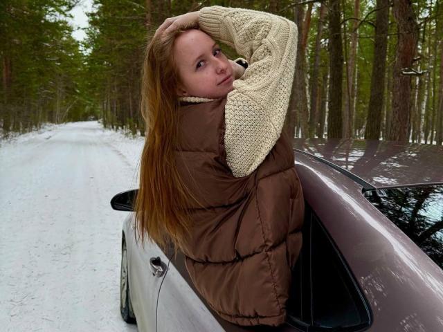 EvangelinaGrubman with long red hair, brown puffy vest, and cream knit sweater, leaning on a brown car, hand in hair, looking back with a slight pout, snowy forest road.