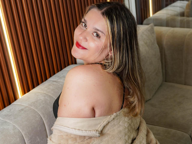 EvelynBlue tem cabelo loiro, batom vermelho e um sorriso adorável. Ela está vestindo uma blusa preta com um casaco bege de ombro caído, que mostra seu ombro e suas costas. Ela está sentada em um sofá..