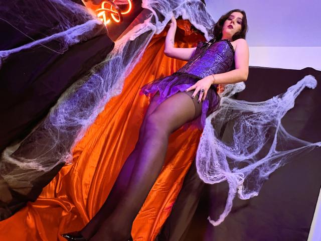 GoddessBar con bustier di pizzo viola, calze nere e un abito corto viola con volant, in piedi tra ragnatele, che guarda verso il basso con labbra rosse, capelli neri e uno sguardo seducente..