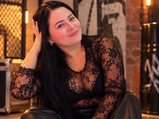 MatureeDesiree with long black hair, wearing a sheer lace top showing her tits, black leather pants, smiling, hand in hair, soft lighting, brick wall in background.