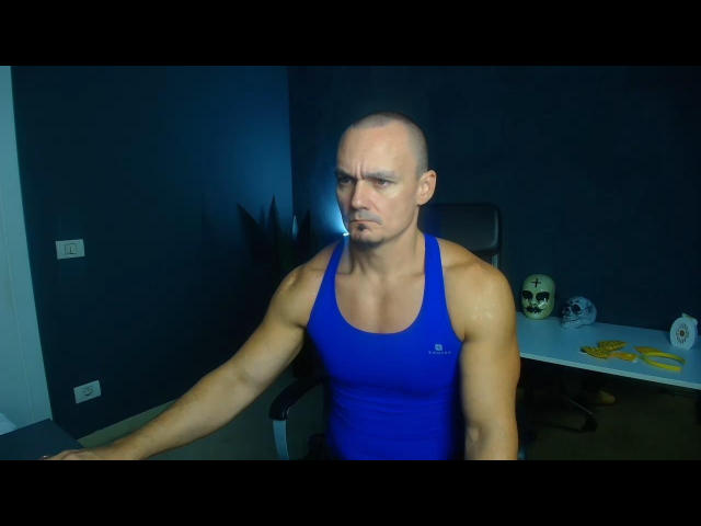 Muscular KaelSage with bald head, blue Under Armour tank, frowning, arms resting on dark chair, in dimly lit room, skull and yellow rope on desk to right.