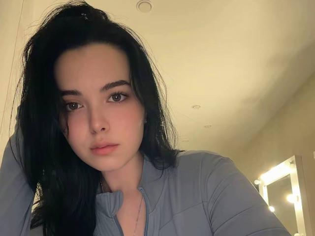 RosalynPullman with black hair, grey shirt, and serious expression, leaning on a surface with her right hand. Her tits are slightly visible under the shirt.