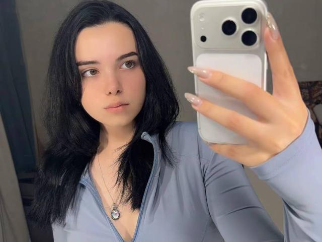 RosalynPullman with black hair, light blue zip-up, holding iPhone, long nails, wearing necklace, taking selfie, neutral expression, pale skin, brown eyes, in mirror selfie.