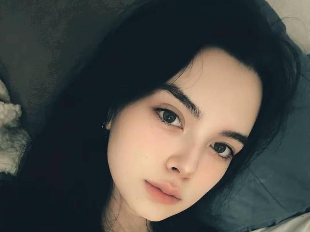 RosalynPullman has pale skin, black hair, and small, pink lips with a slight smile. She has dark eyebrows and green eyes, lying down with a soft, relaxed expression.