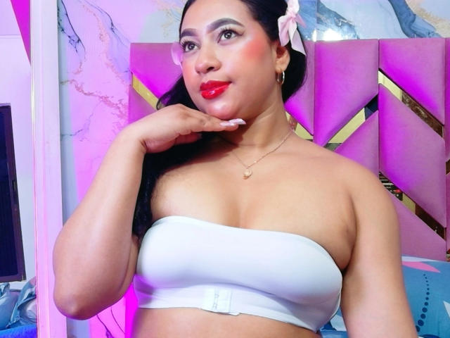 Latina met een medium bruine huid, zwart haar met een roze bloem, een witte top, rode lippenstift, een gouden ketting, die haar kin aanraakt, een paars en roze hoofdeinde, zichtbare borsten, een zelfverzekerde blik..