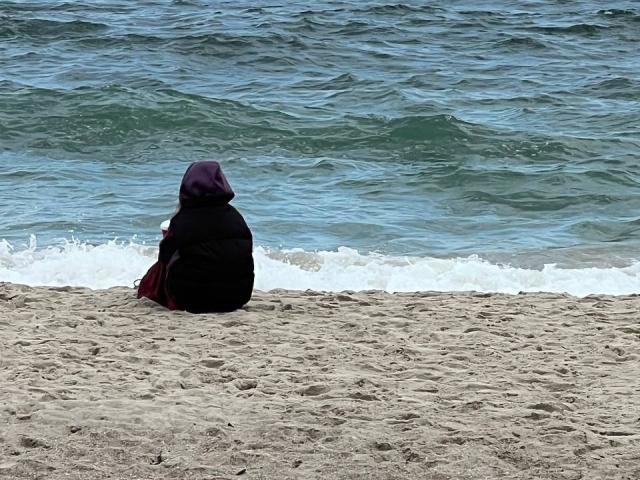 AlexXas sits on a sandy beach, facing the ocean, wearing a black hijab and red dress, with the blue-green water and white waves in the background.