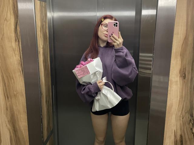 AlexXas with red hair, glasses, purple sweatshirt, black shorts, holding pink flowers in elevator, taking mirror selfie with pink phone, wood-paneled walls.