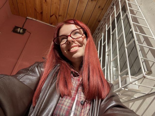 AlexXas with red hair, glasses, smiling, leather jacket, plaid shirt, sunlight on face, wooden ceiling, white metal window, taking a selfie, looking happy, cute, and relaxed.