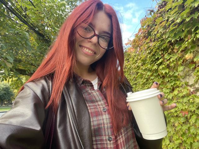 AlexXas with red hair, glasses, black leather jacket, plaid shirt, holding a white coffee cup, smiling, outdoors, green leafy wall, blue sky, and sunlight in the background.