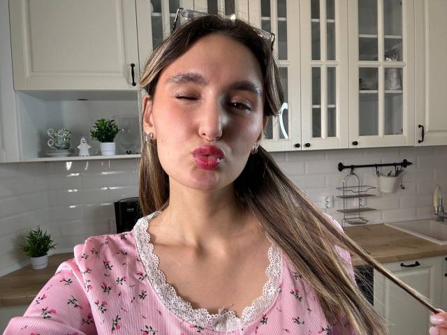 KellyCowling with pouty pink lips, winking, wearing a pink floral shirt with lace, brown hair in ponytail, holding hair, white kitchen cabinets in background, cute and sexy.