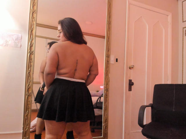 ValerieSynclair is topless with a black skirt, showing her back with a tattoo, standing in front of a mirror, reflecting her butt and long dark hair.