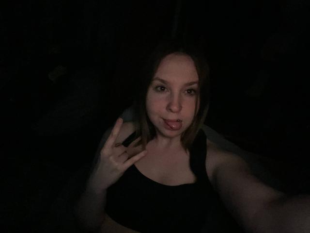 CarmelaBonuz with light skin and brown hair, wearing a black sports bra, making a peace sign with her right hand, mouth slightly open, in a dimly lit setting.