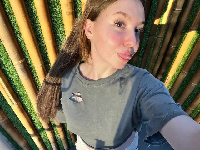 CarmelaBonuz with puffy pink lips, green eyes, and brown hair in a ponytail, wearing a gray tee, standing against bamboo, showing a small tear in her shirt.