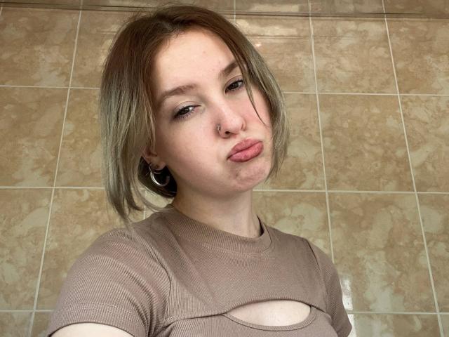 CarmelaBonuz with short brown hair, nose ring, and hoop earrings, pouting lips, wearing a cutout beige top, showing small tits, standing against tiled wall.