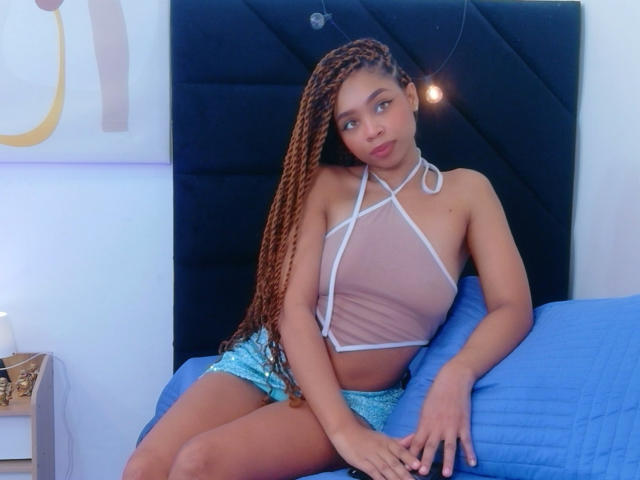 SkinnySamantha, avec ses longs cheveux bruns tressés, portant un haut blanc transparent à encolure américaine et un short bleu pailleté, assise sur un lit bleu, ses petites seins et son joli visage étant visibles..