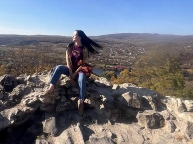 MirandaHott69 with long black hair, wearing a red top and blue jeans, sitting on rocky terrain, smiling, looking up, clear blue sky, mountains in the background.
