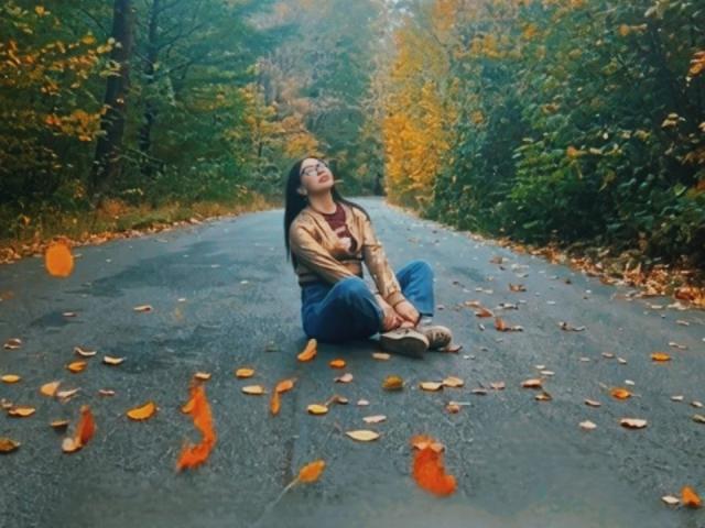 MirandaHott69, sitting on a road with autumn leaves, wearing a brown jacket, blue jeans, and white sneakers, head tilted back, eyes closed, surrounded by falling orange leaves.