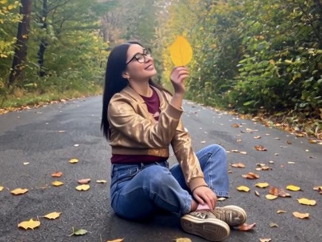 MirandaHott69 sits on a wet road holding a yellow leaf, wearing glasses, a tan jacket, red shirt, blue jeans, and beige sneakers, with autumn leaves scattered around.