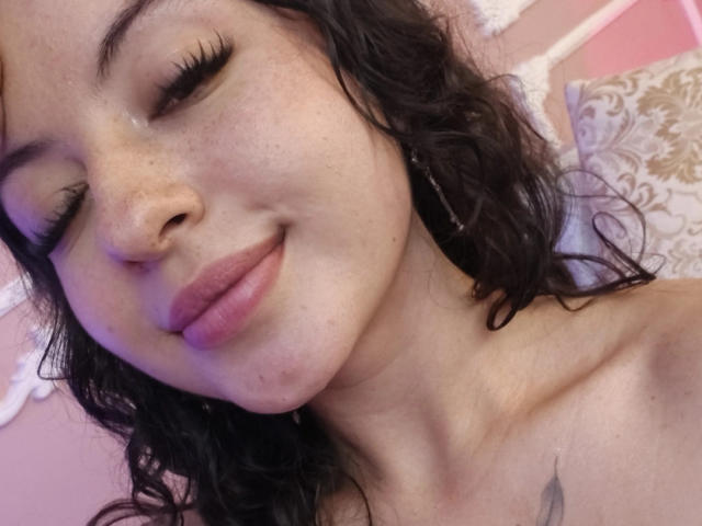 MiiaBennet has curly black hair, light freckles, and is smiling with closed eyes, showing off her pink lips and small tattoo on her chest. Her nipples are visible.