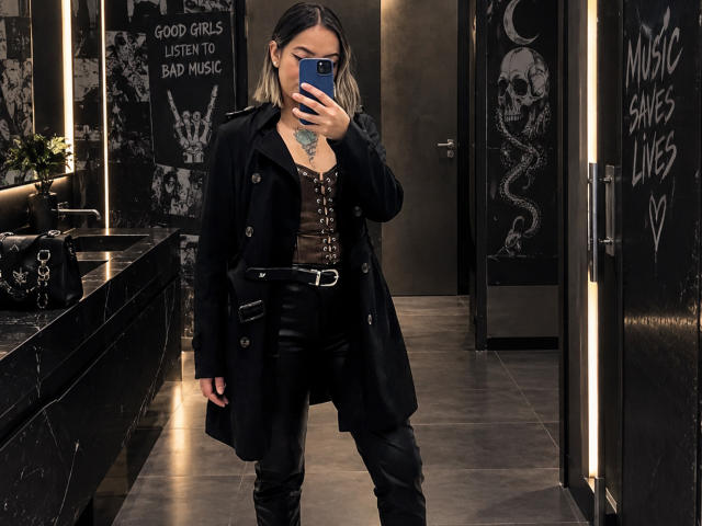 KathyM with black coat, brown top, leather pants, belt, and boots, taking a mirror selfie in a dark, gothic-style bathroom. She has a tattoo on her chest.