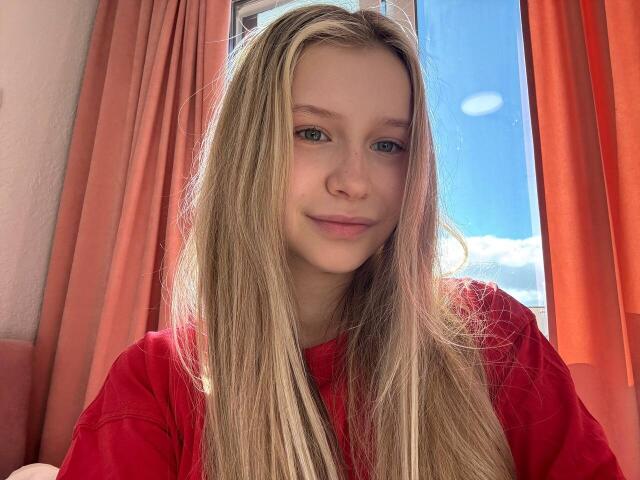 RaeleneRaya has long blonde hair, blue eyes, and a slight smile, wearing a red shirt, with sunlit red curtains in the background. She's cute and looks relaxed.