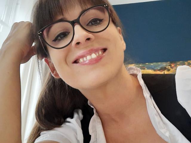 LaLeticia-hot with glasses, smiling, holding her brown hair, wearing a white shirt, showing her teeth, and looking cute with her tits partially visible under the shirt.