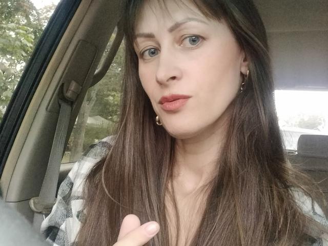 MiraMixi with long brown hair, green eyes, and pink lips, wearing a checkered shirt, inside a car, looking at the camera, with a slight smile and raised finger.