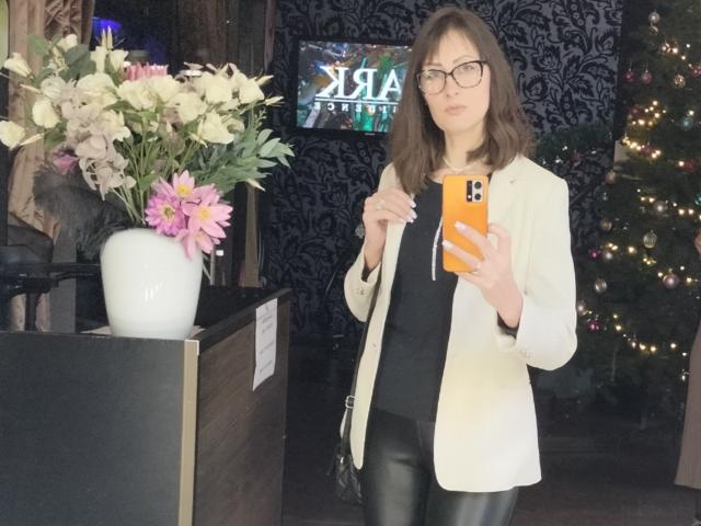 MiraMixi met een bril, die een oranje telefoon vasthoudt, gekleed in een witte blazer, een zwarte top en zwarte leren broek, staat naast een bloevas, voor een versierde kerstboom..