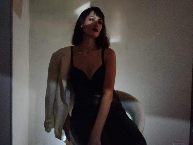 MiraMixi with medium tits, dark brown hair, wearing a black dress and white cardigan, standing in dim light, looking slightly to the side, with shadows on her face.