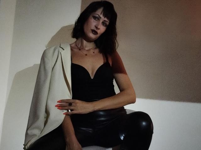 MiraMixi with dark lipstick, black strapless top, white jacket, and black leather pants, sitting, hand on thigh, neon nails, shadowed, looking at camera.