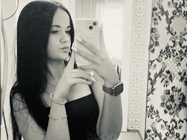 RoxyVesper with long black hair, wearing a black off-shoulder top, holding a phone, taking a mirror selfie, showing her tits, wearing a bracelet, and a watch.