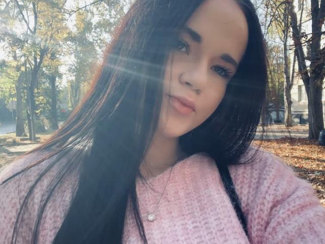 RoxyVesper with long black hair, pink sweater, and subtle makeup, sunlight flares in her face, small heart necklace, soft smile, autumn trees in background, cute, sexy.