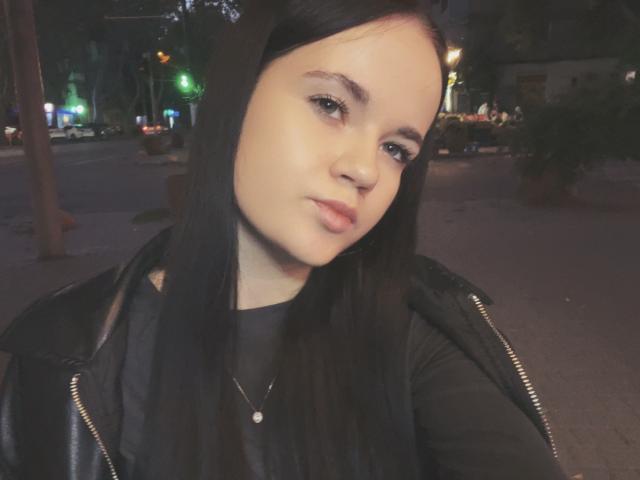RoxyVesper with pale skin, black straight hair, wearing a black leather jacket, black top, and necklace, pouting slightly, in a nighttime urban setting.