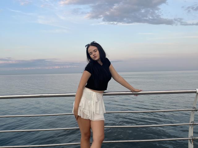 RoxyVesper with black top, white skirt, standing by metal railing, hand on rail, ocean in background, dark hair, sunglasses on head, looking slightly to the side, dusk sky.