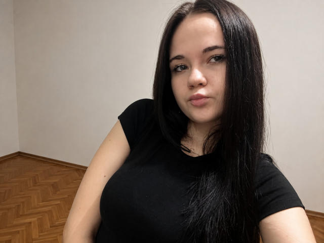 RoxyVesper has long black hair, a pale skin tone, and is wearing a black t-shirt. She's showing off her big boobs in a selfie with a neutral expression.