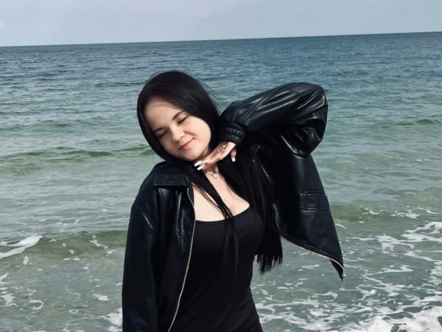 RoxyVesper with black hair, closed eyes, and a smile, wearing a black leather jacket and dress, standing by the ocean, hand on neck, showing her tits subtly.