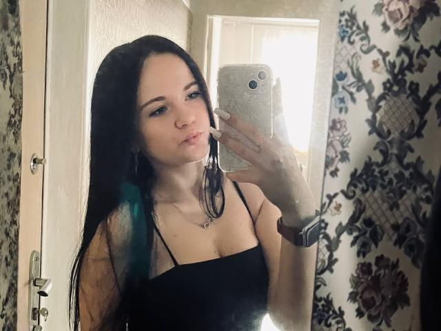 RoxyVesper with long black hair, pouty lips, wearing a black spaghetti strap top, taking a mirror selfie with a silver phone, blue highlights in her hair.