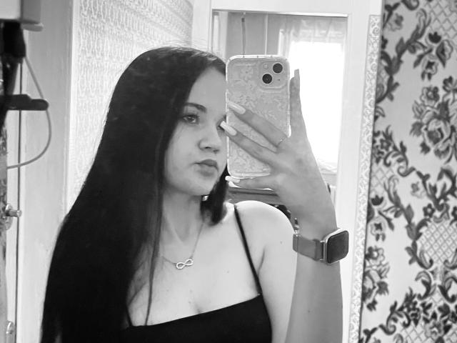 Black-and-white selfie of RoxyVesper with long dark hair, wearing a black spaghetti strap top, holding a phone with a floral case, wearing a necklace and wristwatch.