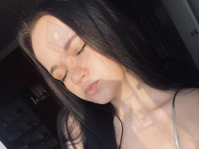 RoxyVesper with closed eyes, long black hair, and pale skin, sunlight casting shadows on her face, wearing a grey strap, slightly pouty lips, soft light, relaxed expression.
