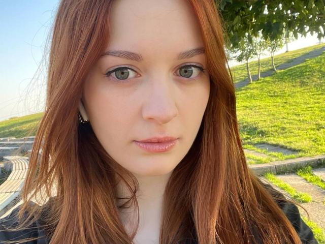 CaritaSchwarzkopf tiene la piel pálida, ojos verdes y cabello rojo. Sus pequeños senos son ligeramente visibles debajo de una camisa negra. Está de pie al aire libre, con una colina cubierta de hierba al fondo..